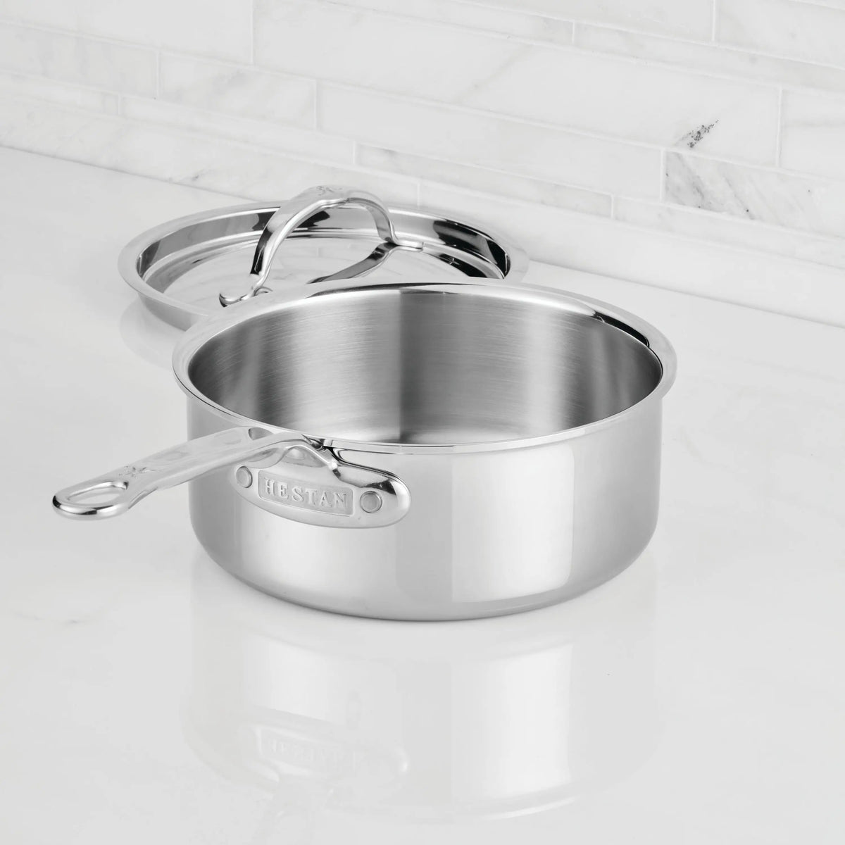 A ProBond® Luxe™ Polished Clad Stainless Steel Saucepan with matching lid rests empty on a white countertop with a white tile backsplash, the lid set slightly askew.