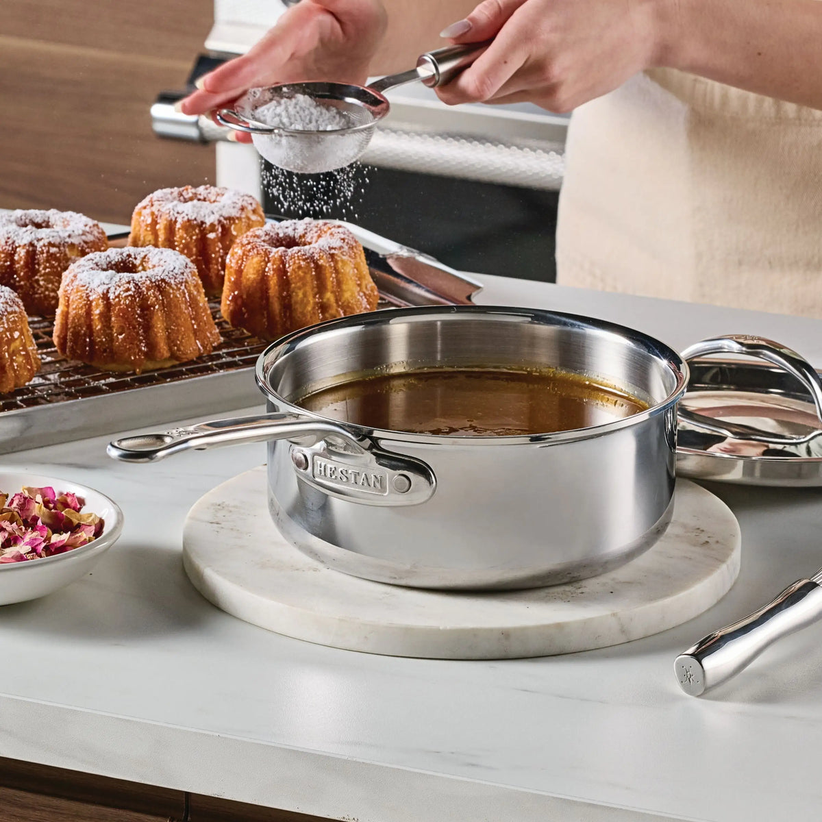 A person dusts powdered sugar over mini bundt cakes on a cooling rack beside a ProBond® Luxe™ Polished Clad Stainless Steel Saucepan with Lid filled with caramel sauce, all arranged on a marble surface.