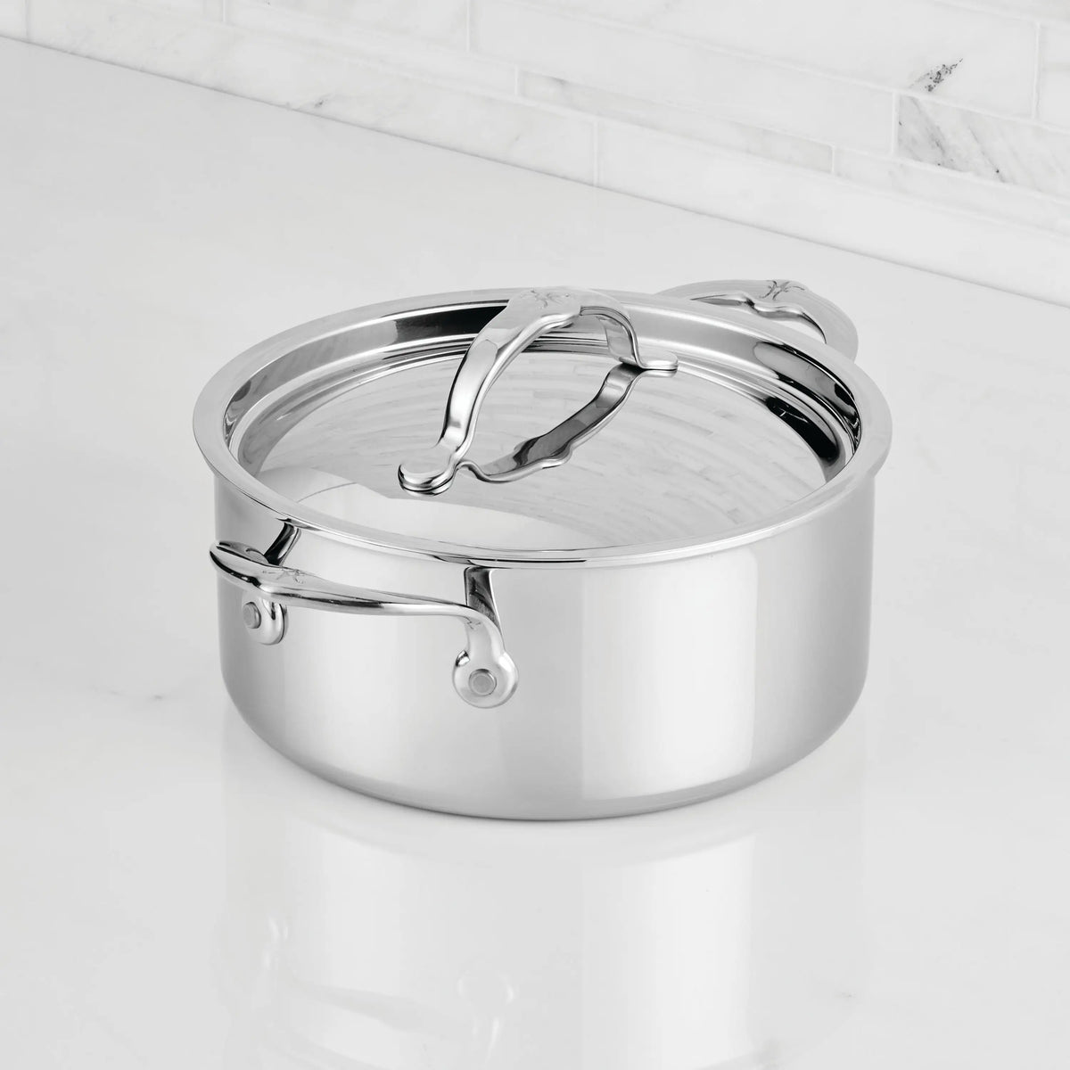 The ProBond® Luxe™ Professional Clad Stainless Steel Soup Pot with Lid, featuring handles and Italian craftsmanship, sits on a white marble countertop against a white tile backsplash.