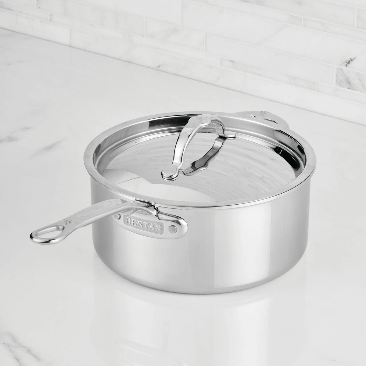 A ProBond® Luxe™ Polished Clad Stainless Steel Saucepan with lid sits on a white marble countertop in front of a light-tiled backsplash, its handle featuring the brand name to highlight premium stainless steel cookware.