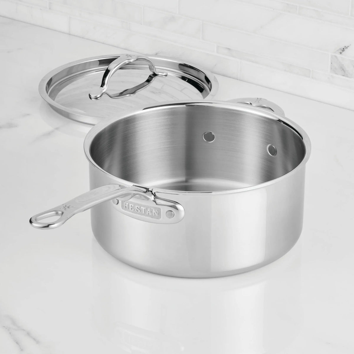 A ProBond® Luxe™ Polished Clad Stainless Steel Saucepan with lid rests on a white marble countertop, the lid propped against the pan and a tiled backsplash visible in the background.