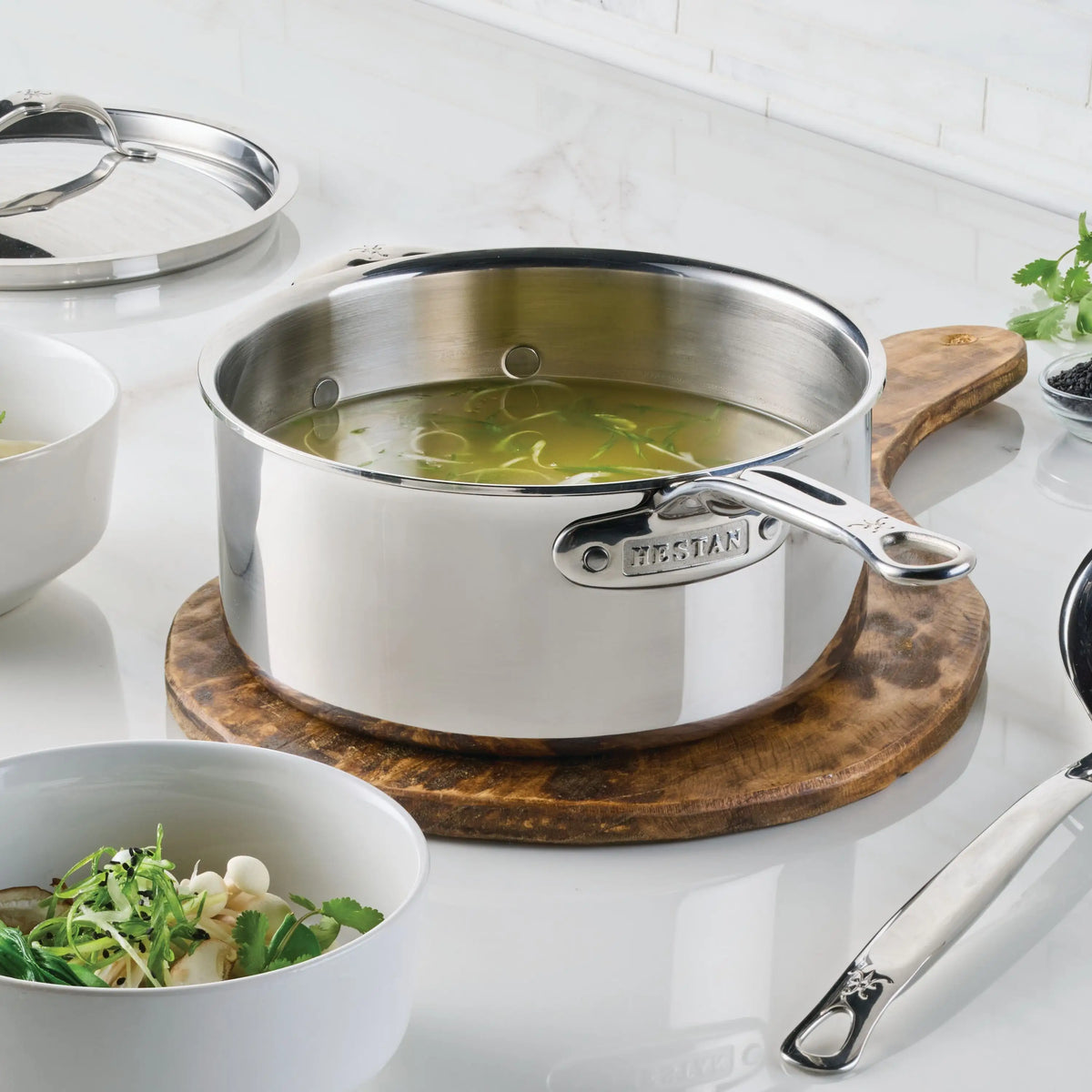 A ProBond® Luxe™ Polished Clad Stainless Steel Saucepan with Lid, filled with soup, rests on a round wooden trivet. Nearby are a bowl of soup with greens and a spoon on a white countertop, plus another pot lid in the background.