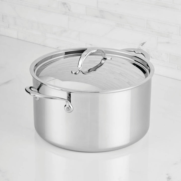 The ProBond® Luxe™ Polished Clad Stainless Steel Stockpot with Lid, featuring two handles and a reflective finish, sits on a white marble countertop with a white tiled backsplash—showcasing refined Italian craftsmanship.