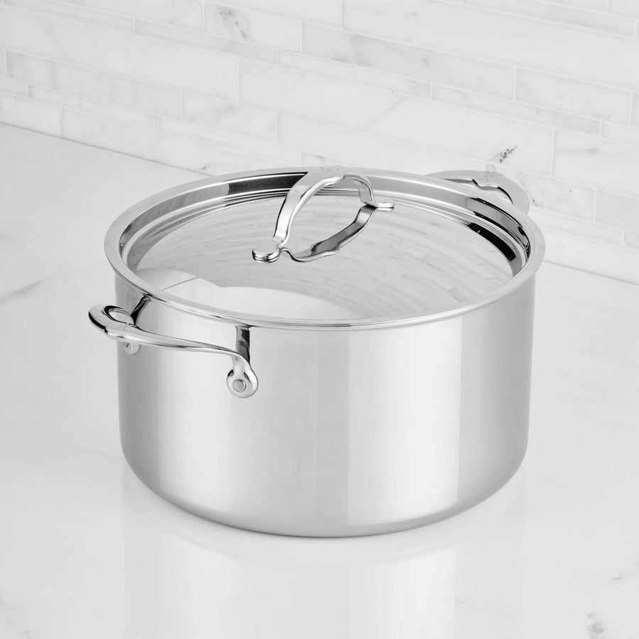 The ProBond® Luxe™ Polished Clad Stainless Steel Stockpot with Lid, featuring two handles and a reflective finish, sits on a white marble countertop with a white tiled backsplash—showcasing refined Italian craftsmanship.