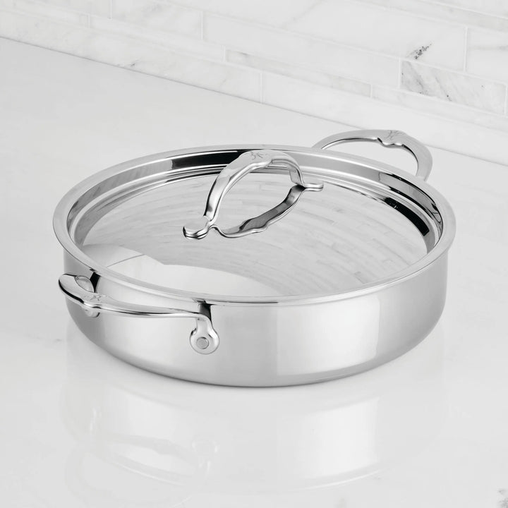 A ProBond® Luxe™ Polished Clad Stainless Steel Rondeaus & Braiser with Lid, featuring curved handles, sits on a white marble countertop against a tiled backsplash.