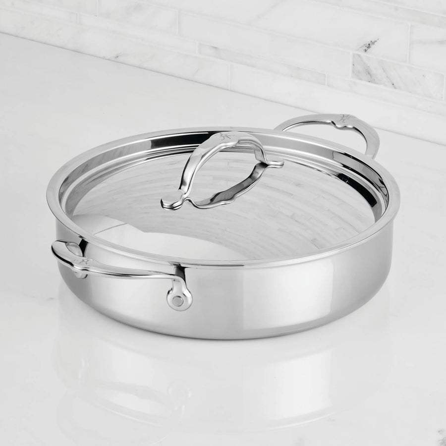 A ProBond® Luxe™ Polished Clad Stainless Steel Rondeaus & Braiser with Lid, featuring curved handles, sits on a white marble countertop against a tiled backsplash.