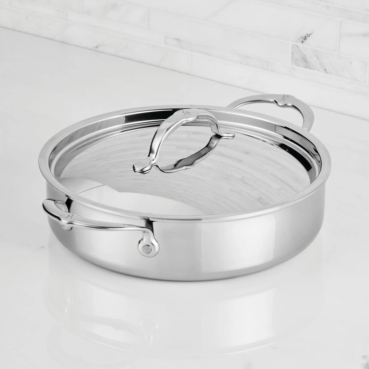 A ProBond® Luxe™ Polished Clad Stainless Steel Rondeaus & Braiser with Lid, featuring curved handles, sits on a white marble countertop against a tiled backsplash.