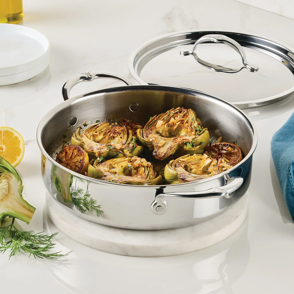 A ProBond® Luxe™ Polished Clad Stainless Steel Braiser with Lid, 3.5-Quart, is filled with roasted artichoke halves on a white surface. The lid rests nearby beside a blue napkin, cut lemon, and fresh herbs.