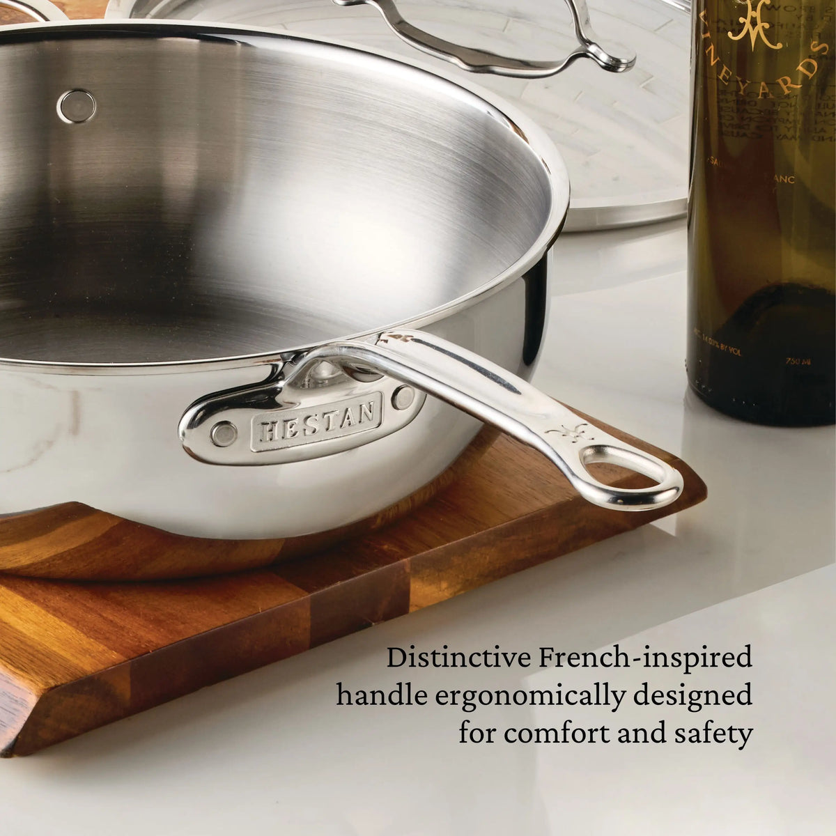 A ProBond® Luxe™ Professional Clad Stainless Steel Essential Pan with Lid rests on a wooden trivet beside a green glass wine bottle. Text reads, “Professional clad stainless steel cookware designed for comfort and safety.”.
