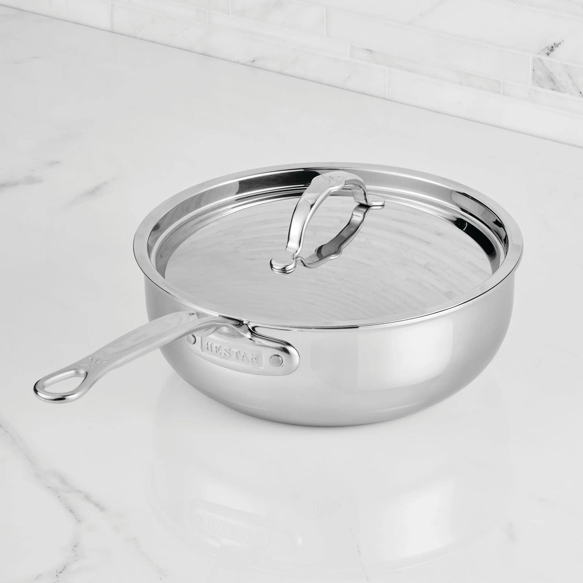The ProBond® Luxe™ Professional Clad Stainless Steel Essential Pan with Lid features a sleek, reflective finish and long handle, resting securely on a white marble countertop.