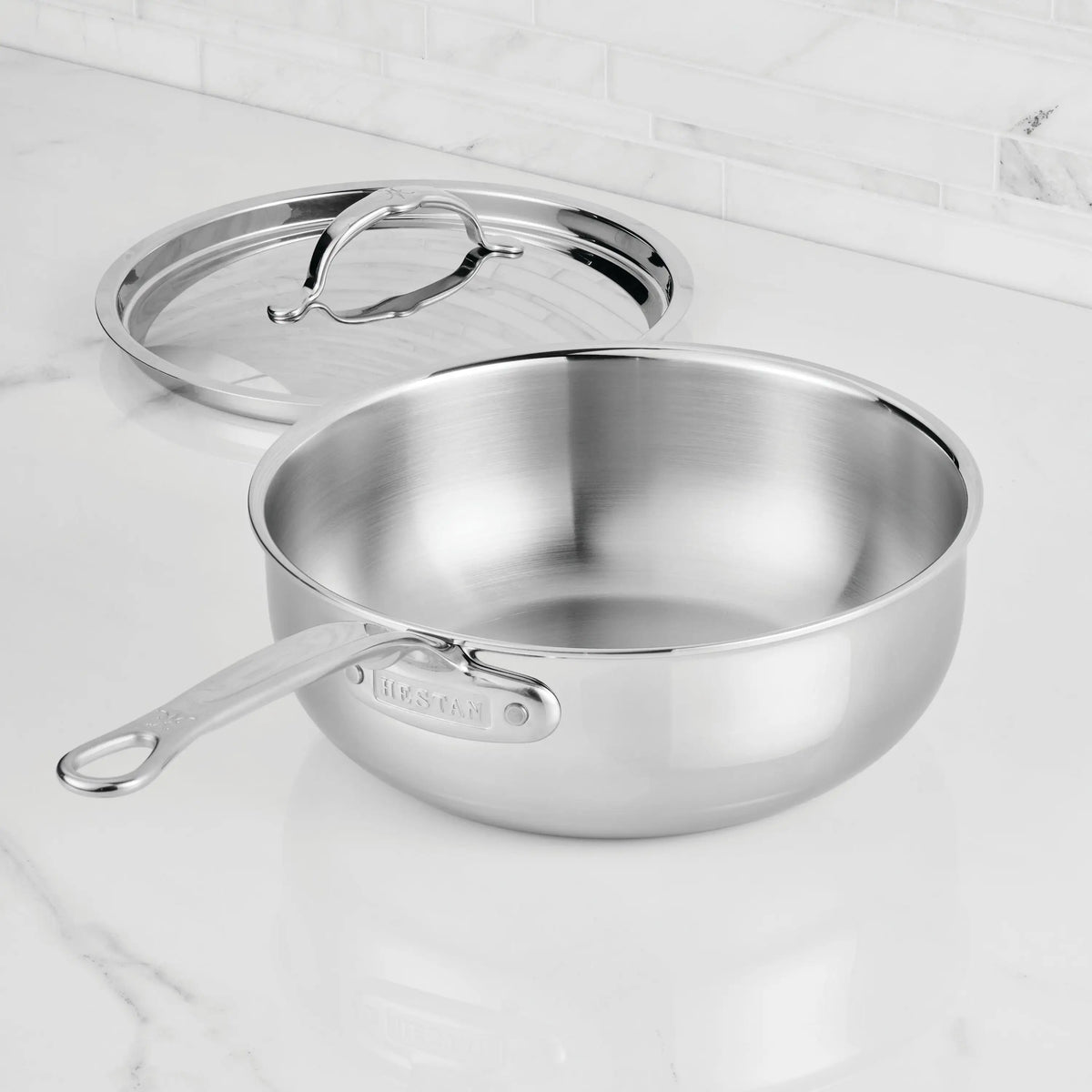 A ProBond® Luxe™ Professional Clad Stainless Steel Essential Pan with Lid sits on a white marble countertop, highlighting its Italian craftsmanship and modern design.