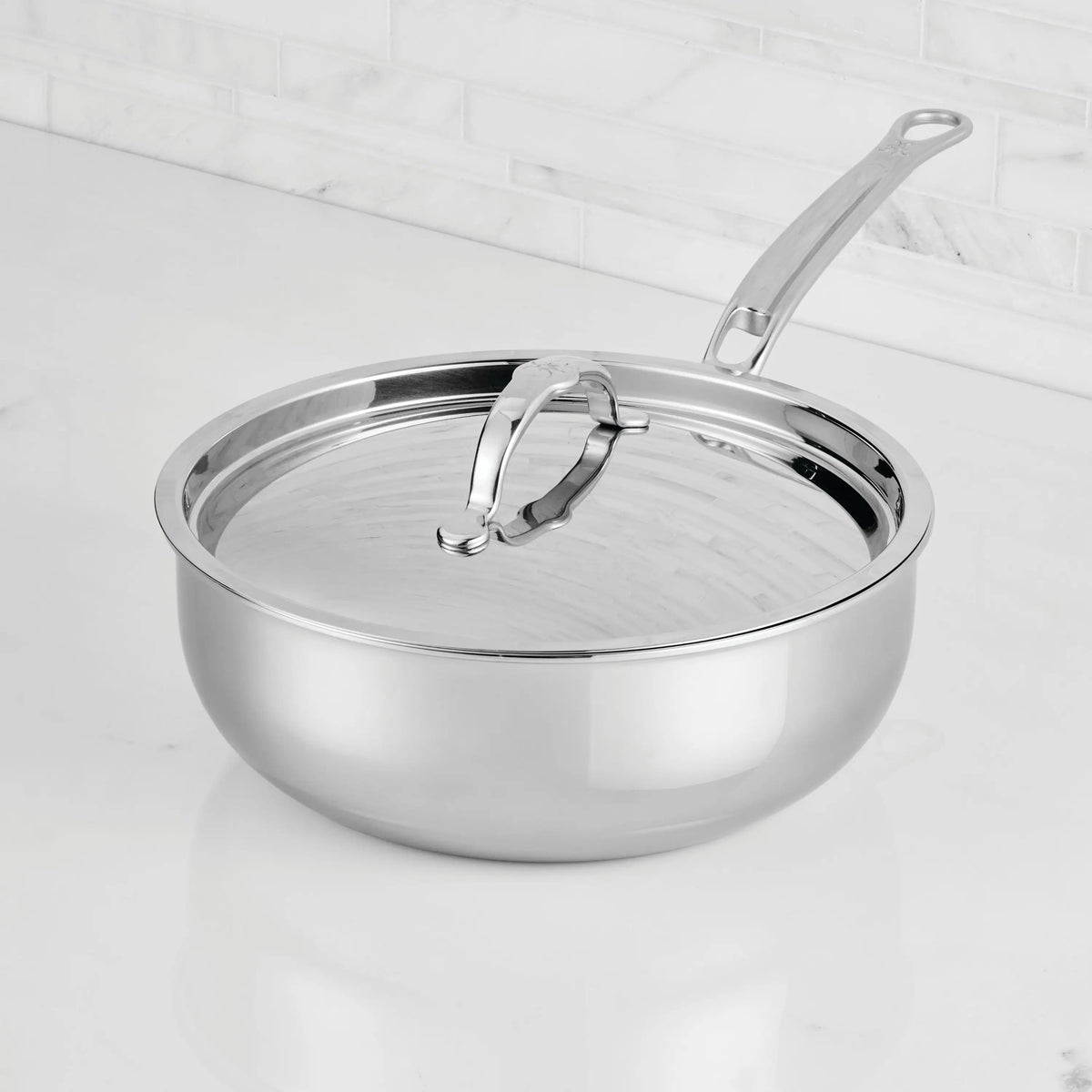 A ProBond® Luxe™ Professional Clad Stainless Steel Essential Pan with Lid rests on a white marble kitchen countertop, with a tiled backsplash in the background.