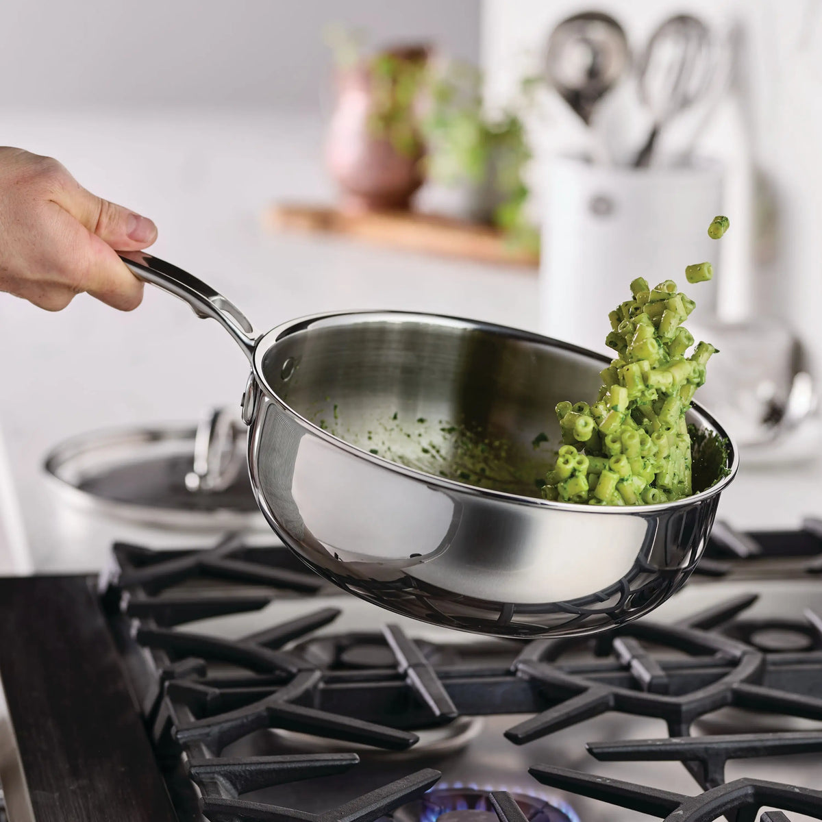 A hand holds a ProBond® Luxe™ Professional Clad Stainless Steel Essential Pan with Lid over a gas stove, flipping chopped green vegetables in mid-air. A glass lid rests nearby on the countertop, with a softly blurred kitchen background.