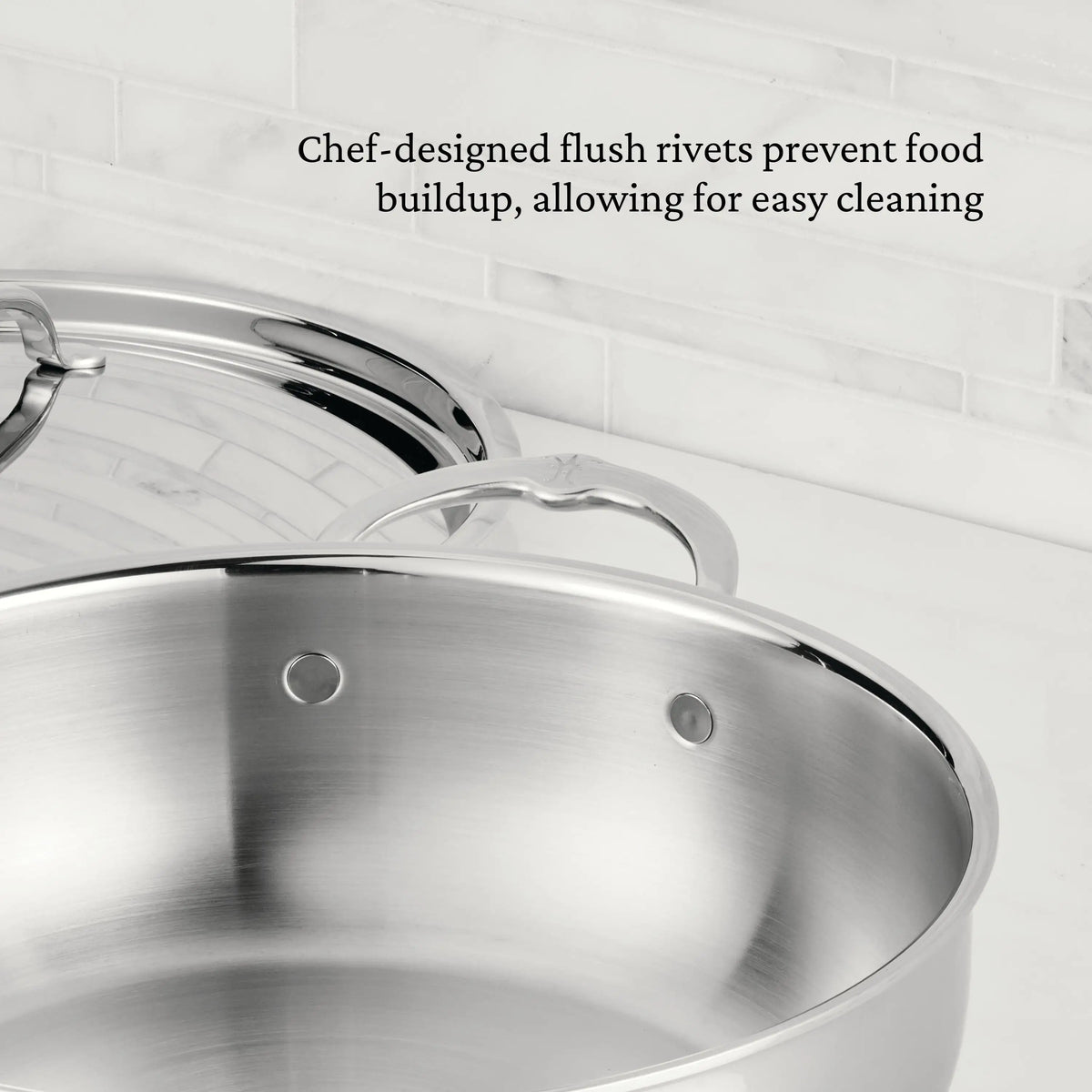 A ProBond® Luxe™ Professional Clad Stainless Steel Essential Pan with Lid sits on a white countertop. Chef-designed, flush rivets inspired by Italian craftsmanship prevent food buildup for effortless maintenance and easy cleaning.