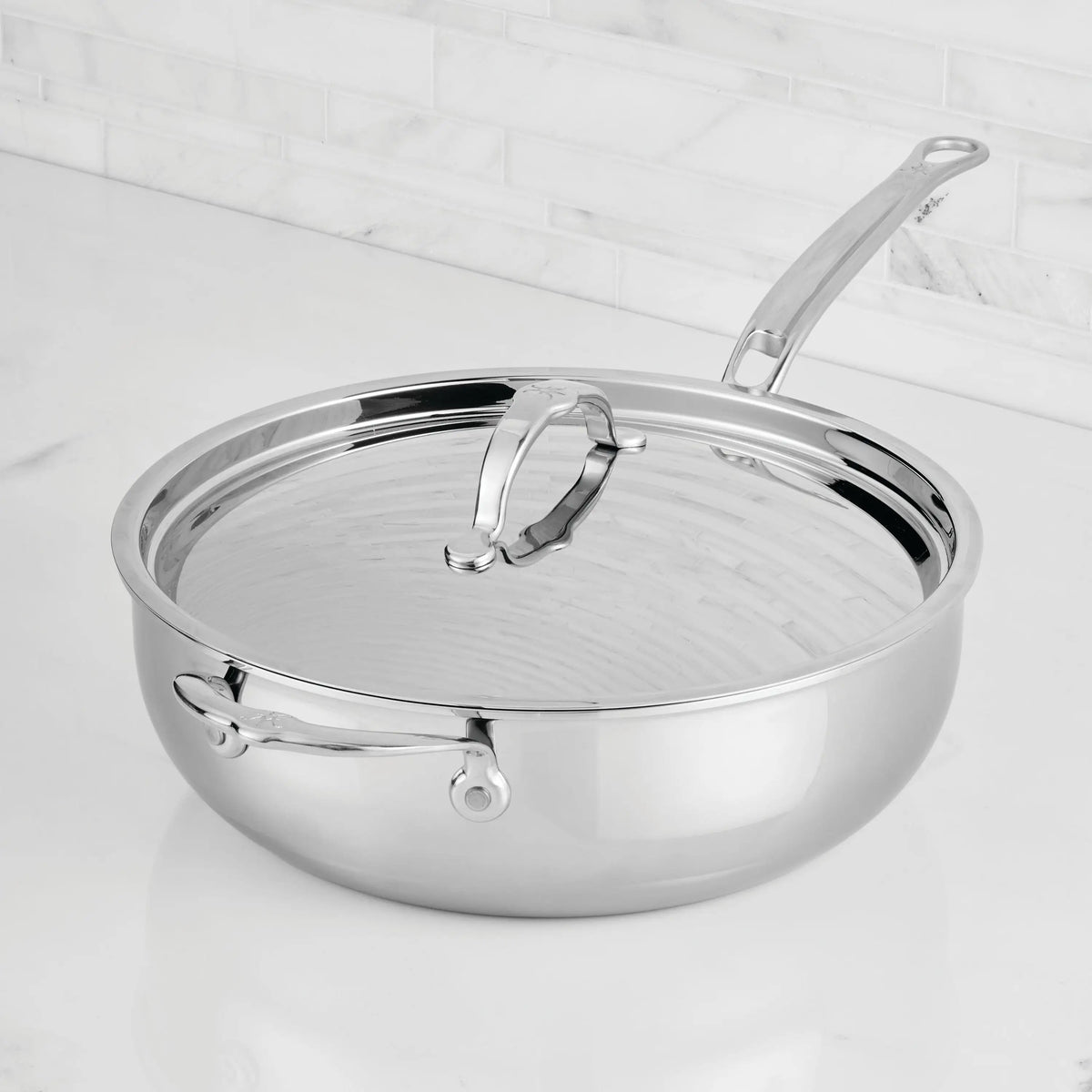 A ProBond® Luxe™ Professional Clad Stainless Steel Essential Pan with Lid and dual handles rests on a white marble counter, set against a white tiled backsplash and showcasing premium Italian-inspired craftsmanship.