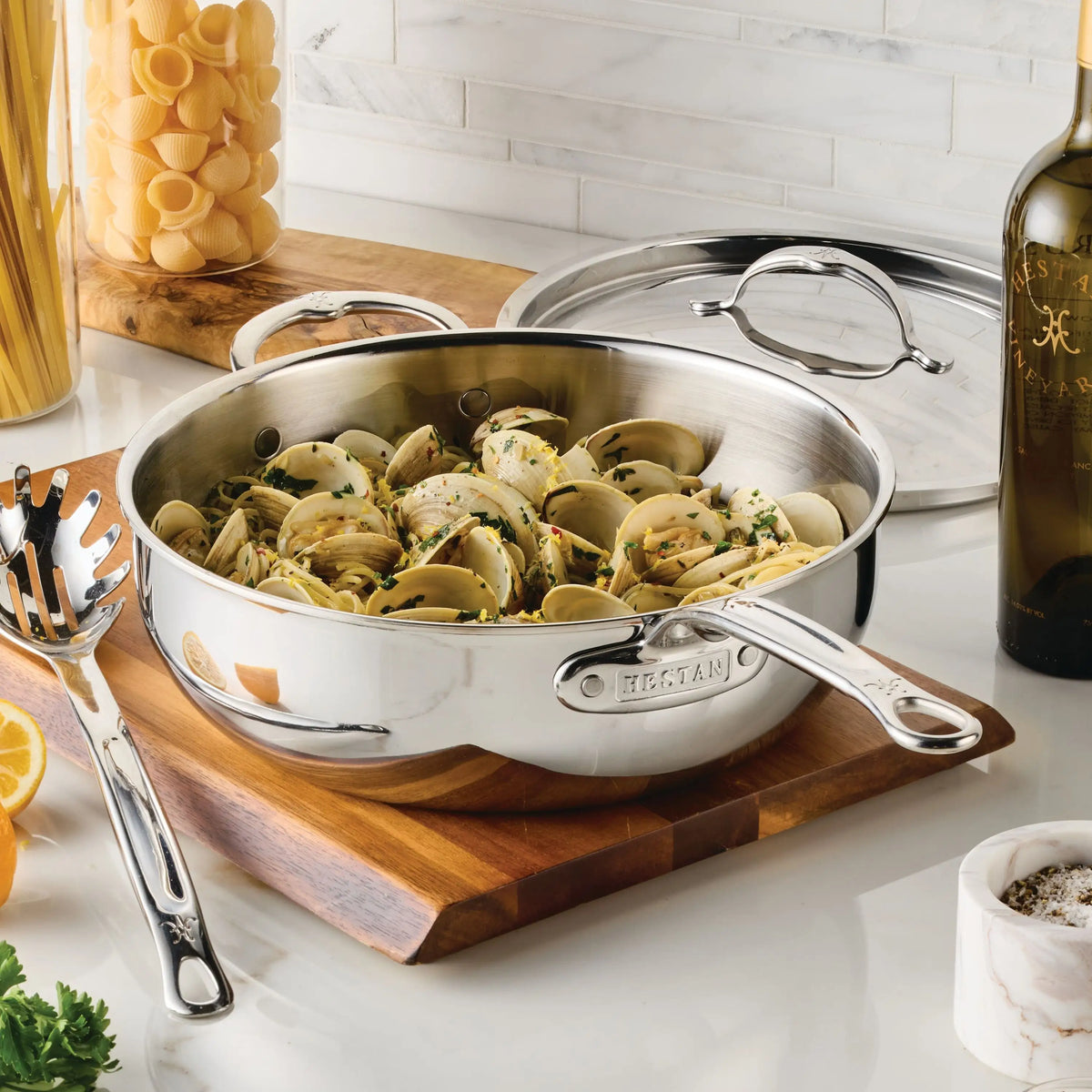 A ProBond® Luxe™ Polished Clad Stainless Steel Signature Set pan brimming with clams and pasta sits on a cutting board, surrounded by lemon halves, fresh herbs, uncooked pasta jar, wine bottle, and a spaghetti server.
