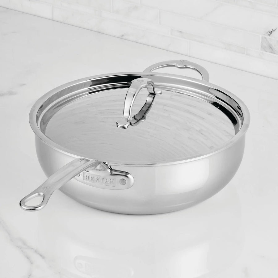 A ProBond® Luxe™ Professional Clad Stainless Steel Essential Pan with Lid rests on a white marble countertop in a kitchen with a white tile backsplash, showcasing premium craftsmanship and style.