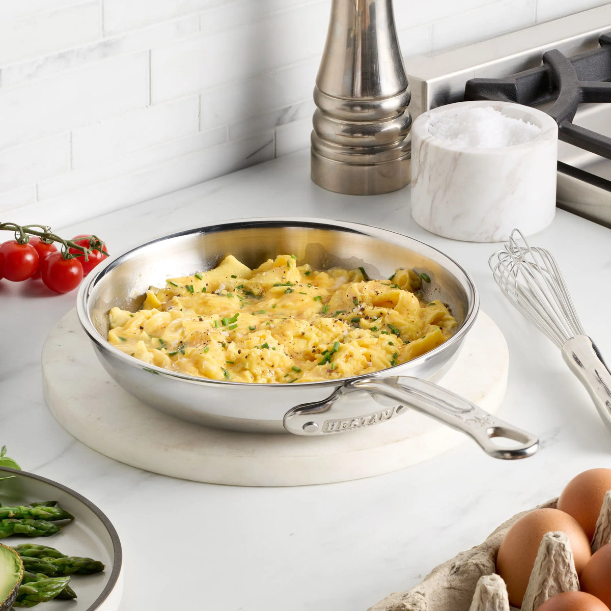 A ProBond® Luxe™ Polished Clad Stainless Steel Skillet with scrambled eggs rests on a marble trivet, surrounded by a whisk, eggs, asparagus, grape tomatoes, salt container, and pepper grinder.