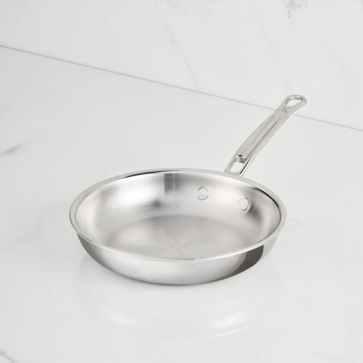A ProBond Luxe® Polished Clad Stainless Steel Skillet with a metal handle sits on a white marble countertop. The induction-suitable skillet is empty and clean, angled slightly to the left.