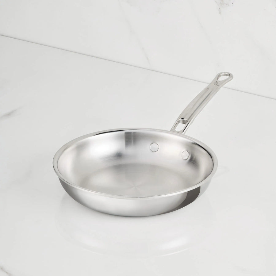 A ProBond Luxe® Polished Clad Stainless Steel Skillet with a metal handle sits on a white marble countertop. The induction-suitable skillet is empty and clean, angled slightly to the left.