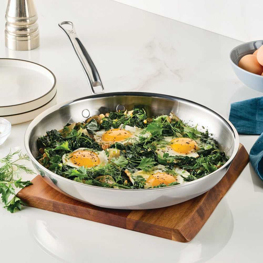 A ProBond Luxe® Polished Clad Stainless Steel Skillet filled with four sunny-side up eggs, cooked alongside greens and herbs, rests on a wooden cutting board atop a white countertop. Surrounding it are plates, a towel, fresh eggs, and a pepper mill.
