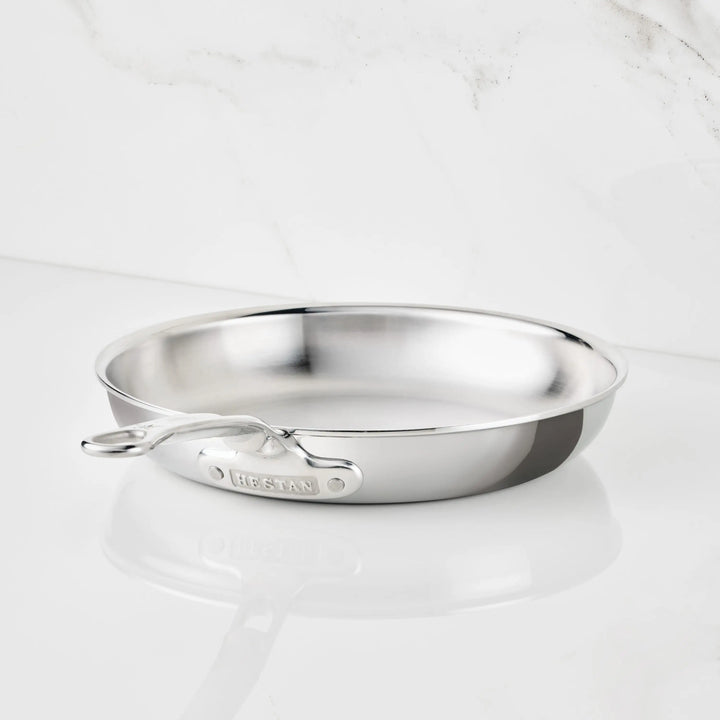 A Polished Clad Stainless Steel Skillet from ProBond Luxe® with a long handle rests on a white marble surface. The tri-ply skillet is empty and gleams as it catches the light, with the brand name ProBond Luxe® visible on the handle.