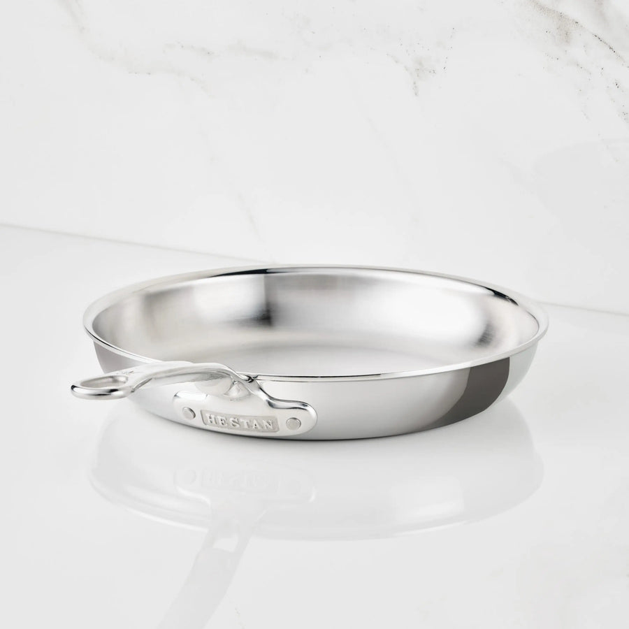 A Polished Clad Stainless Steel Skillet from ProBond Luxe® with a long handle rests on a white marble surface. The tri-ply skillet is empty and gleams as it catches the light, with the brand name ProBond Luxe® visible on the handle.
