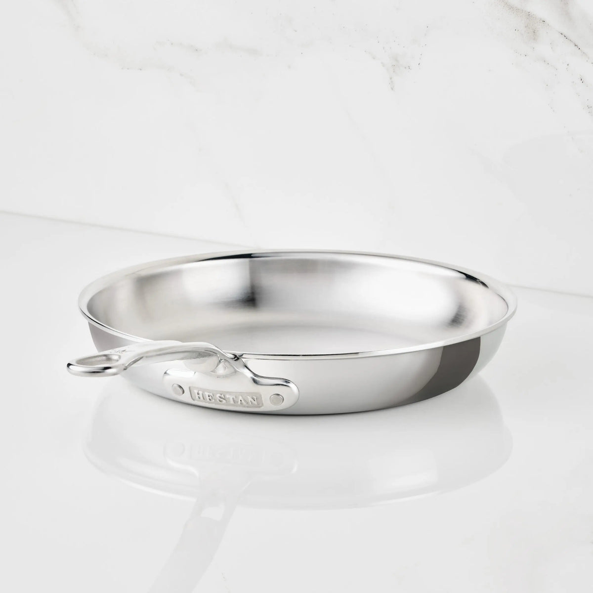 A Polished Clad Stainless Steel Skillet from ProBond Luxe® with a long handle rests on a white marble surface. The tri-ply skillet is empty and gleams as it catches the light, with the brand name ProBond Luxe® visible on the handle.