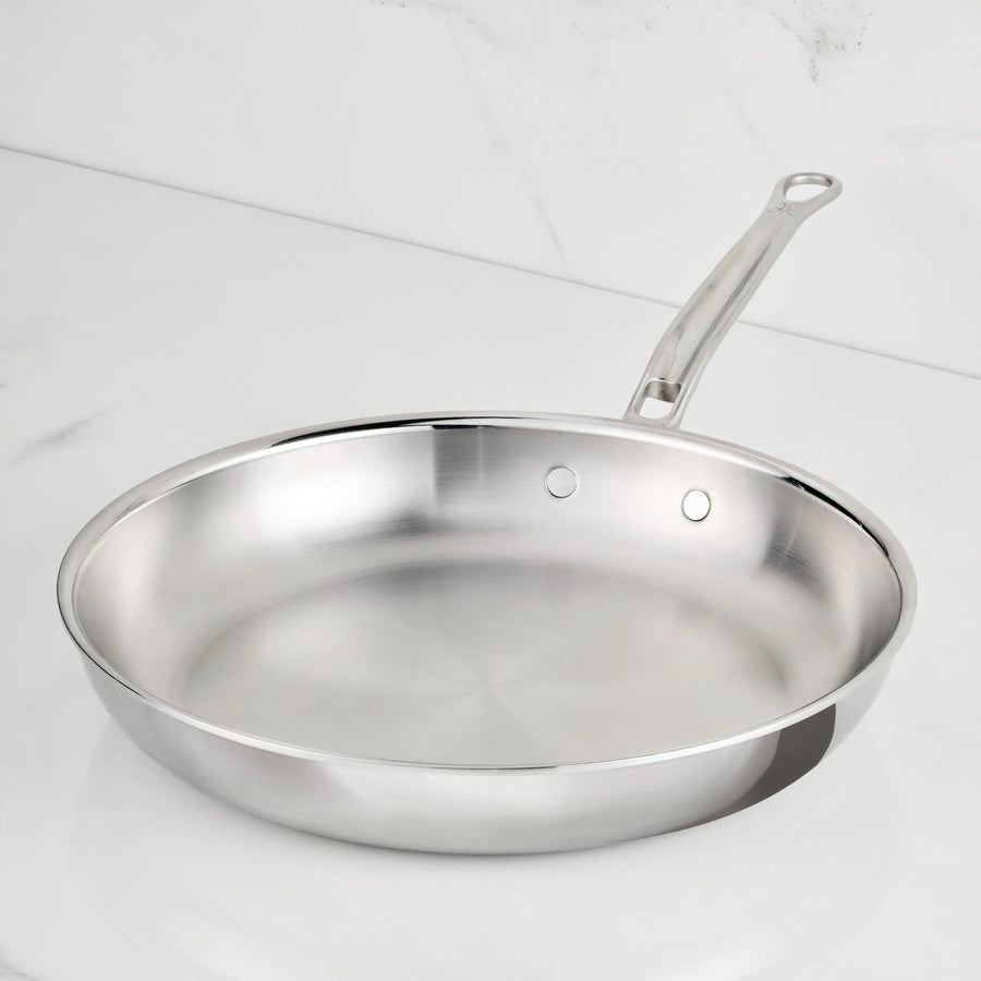 A ProBond Luxe® Polished Clad Stainless Steel Skillet with a long handle rests on a white marble countertop. This skillet is clean, empty, and features a smooth, reflective surface with tri-ply clad construction that makes it induction-suitable for modern kitchens.