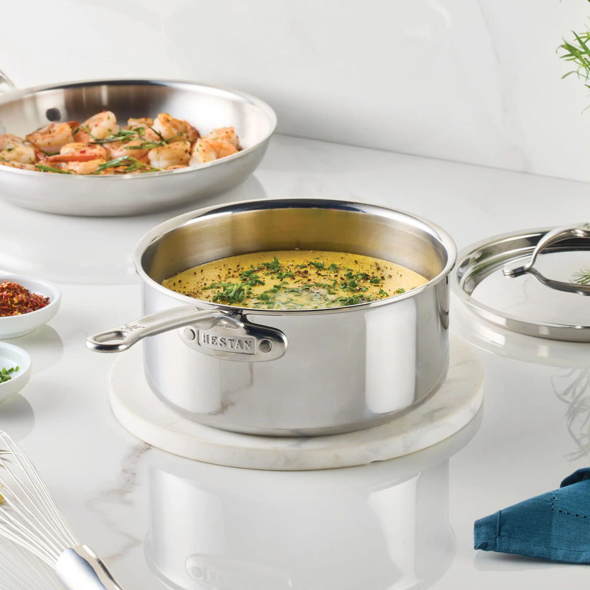 A ProBond® Luxe™ Polished Clad Stainless Steel Saucepan with Lid, filled with yellow soup and herbs, sits on a marble trivet. Behind it, a sauté pan holds shrimp amid kitchen tools and bowls on the white countertop.