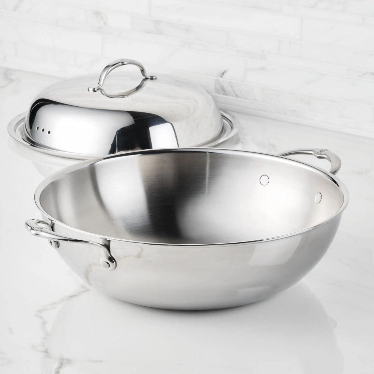 A 14-inch ProBond® Luxe™ Polished Clad Stainless Steel Wok with Dome Lid sits on a marble countertop, featuring professional clad construction and a Hestan ProCore aluminum core, standing out against white marble tiles.