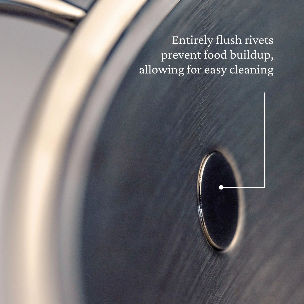 Close-up of a metal surface with a flush rivet on the ProBond® Luxe™ Polished Clad Stainless Steel Wok with Dome Lid, 14-Inch. Text: Flush rivets prevent food buildup for easy cleaning. An arrow highlights the rivet.