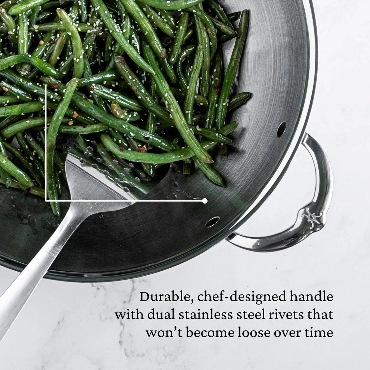 A metal slotted spatula rests in a Polished Clad Stainless Steel Wok with Dome Lid, 14-Inch by ProBond® Luxe™, filled with green beans. The wok’s shiny curved handle has dual stainless steel rivets for lasting durability.