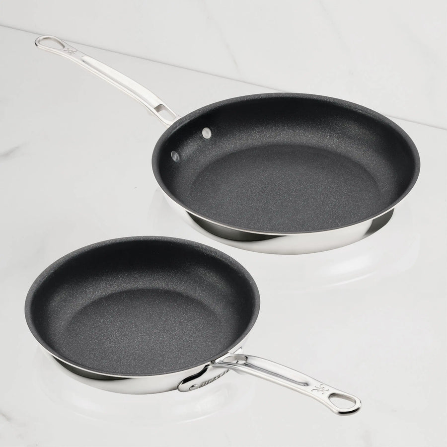 Two ProBond® Luxe™ Polished Clad Stainless Steel Skillets with TITUM® Nonstick, one larger than the other, are displayed on white marble. Their premium nonstick interiors and metal handles combine style and function.
