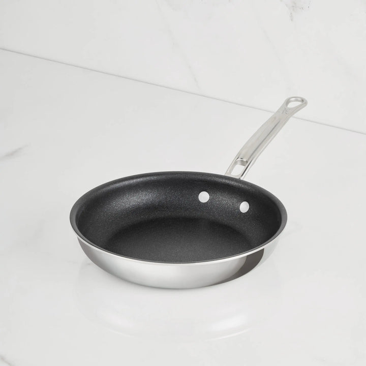 A ProBond Luxe® Polished Clad Stainless Steel Skillet with TITUM® Nonstick, featuring a sleek metal handle and crafted with exquisite Italian craftsmanship, sits on a white marble surface with a white marble background.