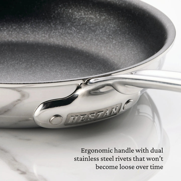 Close-up of a ProBond® Luxe™ Polished Clad Stainless Steel Skillet with TITUM® Nonstick, featuring an ergonomic handle secured by two rivets and the ProBond® Luxe™ logo near the handle, shown on a marble countertop. Text highlights handle features.