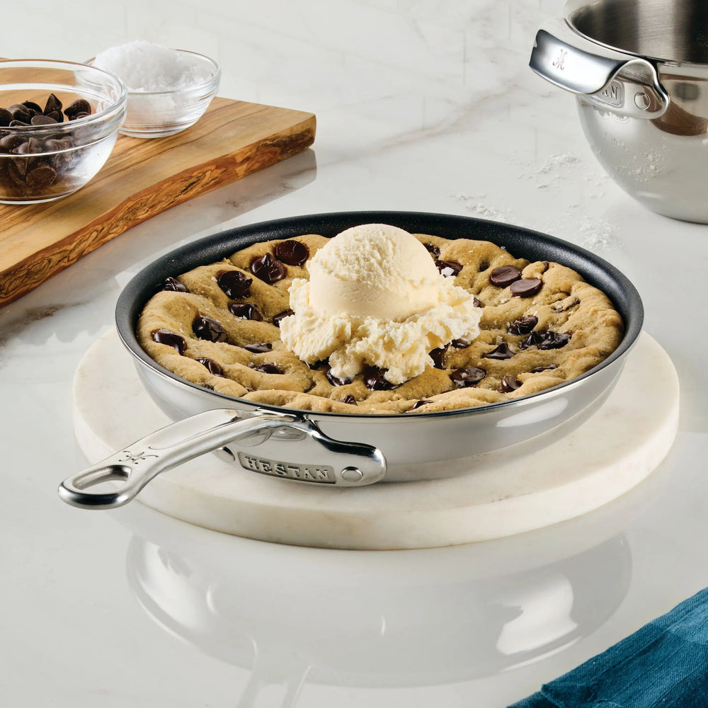 A skillet cookie topped with vanilla ice cream sits on a round marble board, with ProBond® Luxe™ Polished Clad Stainless Steel Skillets with TITUM® Nonstick and baking utensils visible in the background on a white countertop.