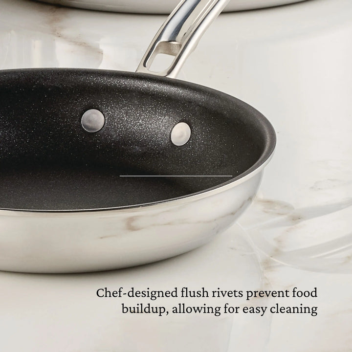 A ProBond® Luxe™ Polished Clad Stainless Steel Skillet with TITUM® Nonstick and two flush rivets sits on a marble countertop. Text below notes that flush rivets prevent food buildup for easy cleaning.