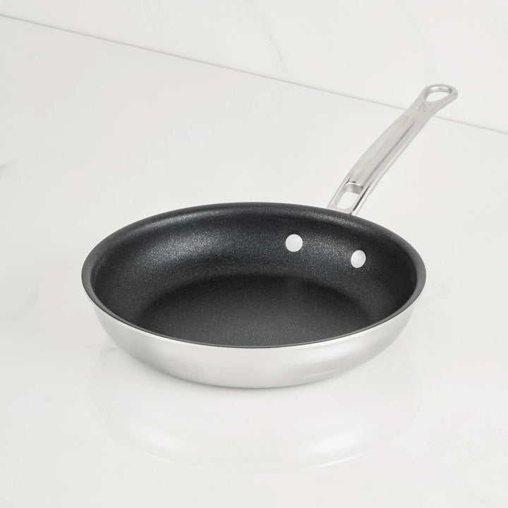 A ProBond® Luxe™ Polished Clad Stainless Steel Skillet with TITUM® Nonstick black interior sits on a white countertop, featuring a long riveted metal handle; its part of premium stainless steel cookware.