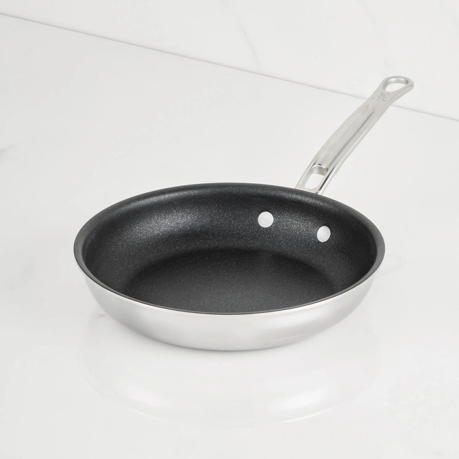 A ProBond® Luxe™ Polished Clad Stainless Steel Skillet with TITUM® Nonstick black interior sits on a white countertop, featuring a long riveted metal handle; its part of premium stainless steel cookware.