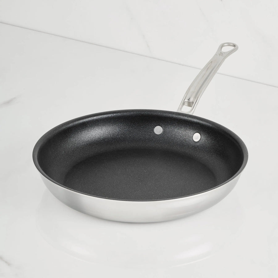 A ProBond® Luxe™ Polished Clad Stainless Steel Skillet with TITUM® Nonstick interior and a metal handle sits on a white marble countertop, backed by a matching marble wall.