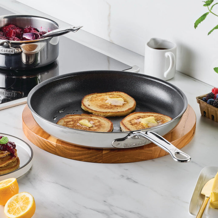 Three pancakes with melting butter sizzle in a ProBond® Luxe™ Polished Clad Stainless Steel Skillet with TITUM® Nonstick on a wooden board, surrounded by berries, cookware, a pitcher, lemon, and a plate with pancake and fork on marble.