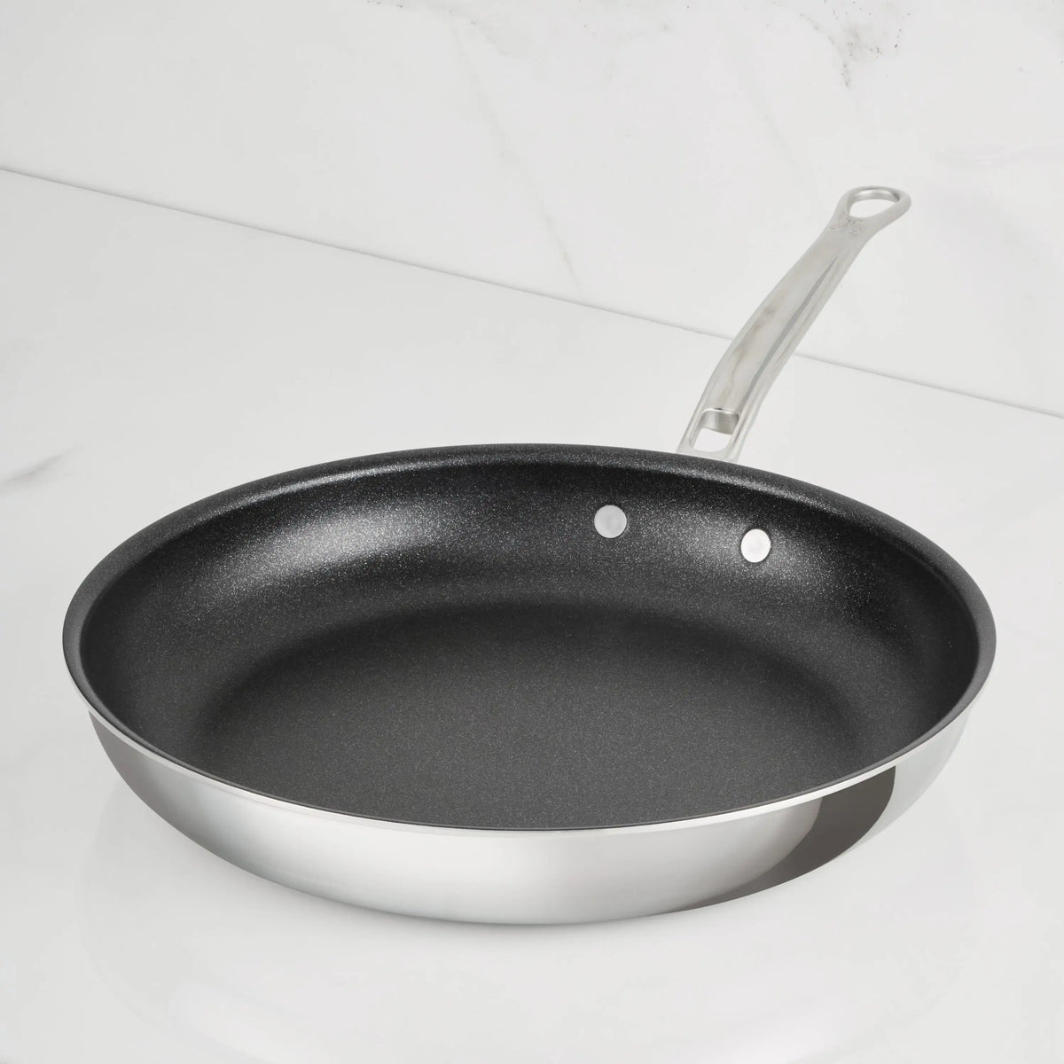 A ProBond Luxe® Polished Clad Stainless Steel Skillet with a TITUM® Nonstick interior and a metal handle sits on a white, marble-like countertop.