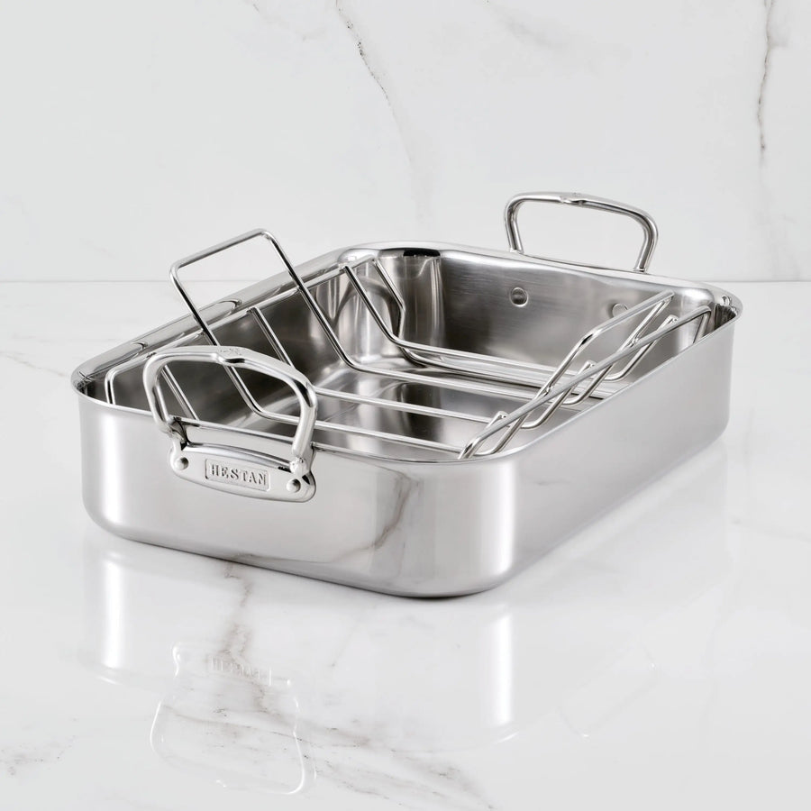 The Hestan Provisions Polished Stainless Steel Roaster, featuring two sturdy handles and a removable rack, sits oven-safe on a white marble surface.