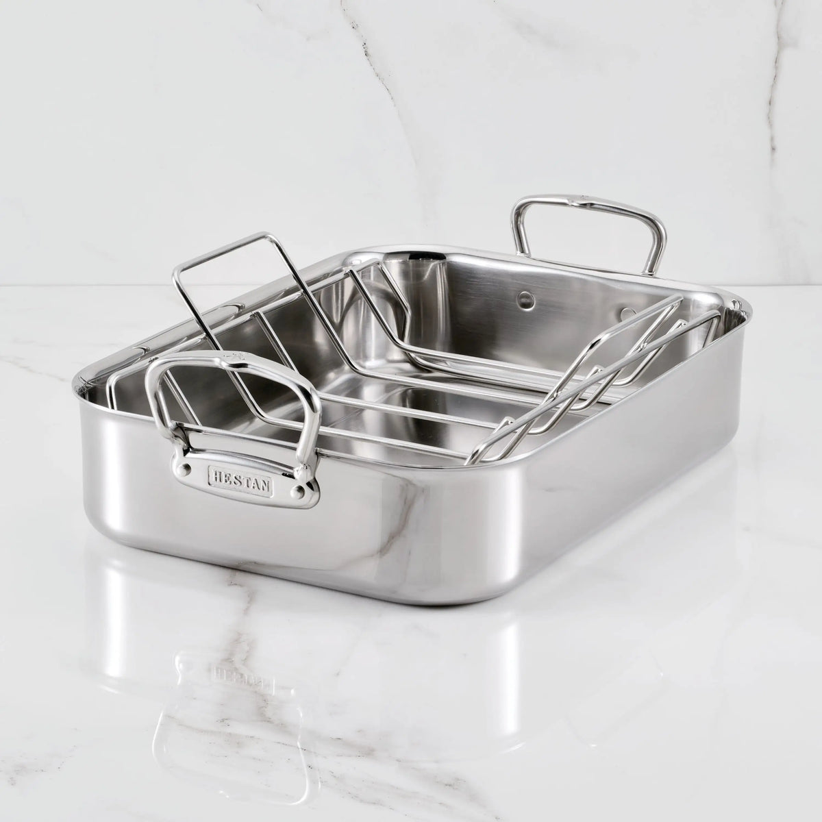 The Hestan Provisions Polished Stainless Steel Roaster, featuring two sturdy handles and a removable rack, sits oven-safe on a white marble surface.