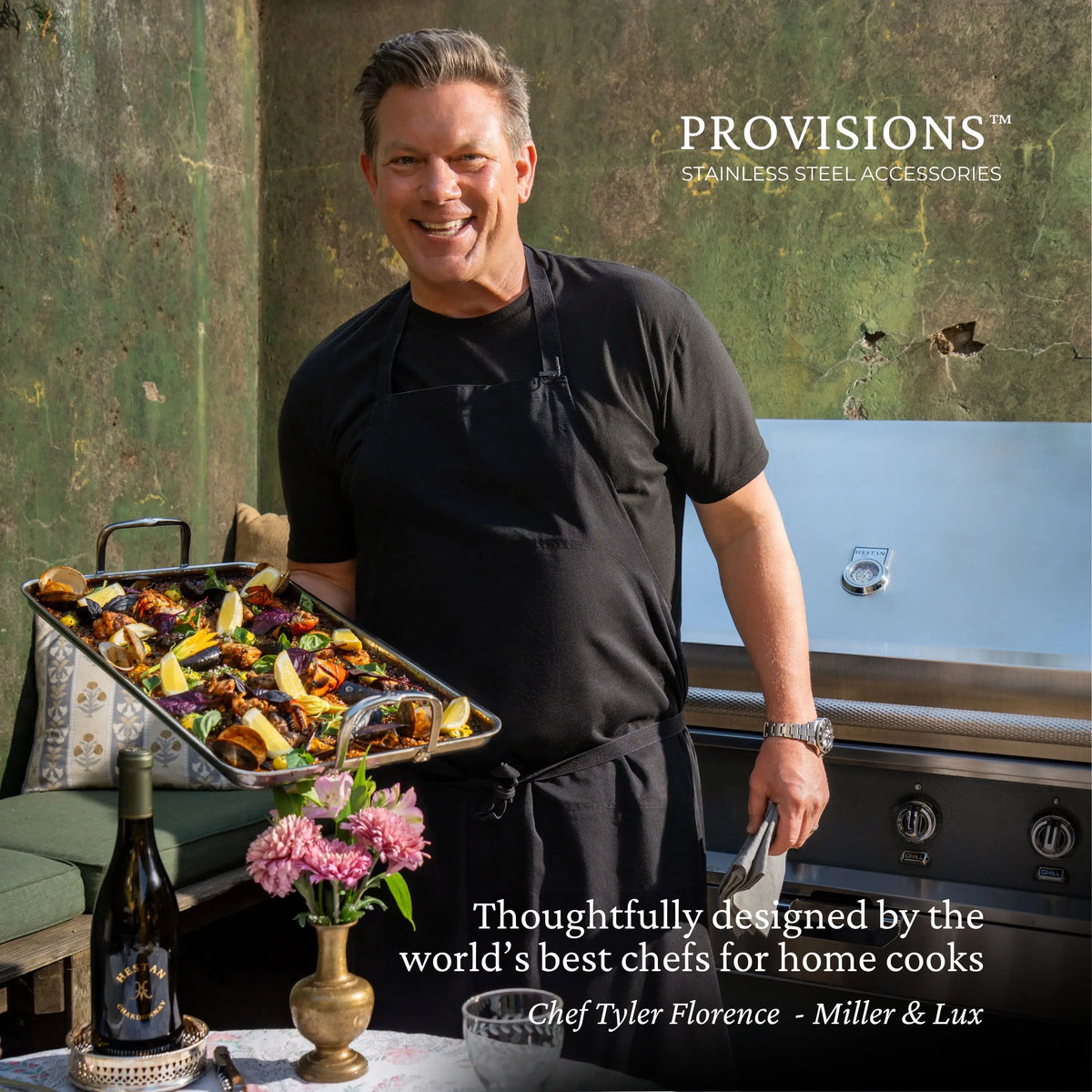 A man in a black apron smiles, holding roasted food by an outdoor grill. On a nearby table are wine, pink flowers, and text highlighting the NanoBond Titanium x Tyler Florence 41-Piece Cookware Set accessories.