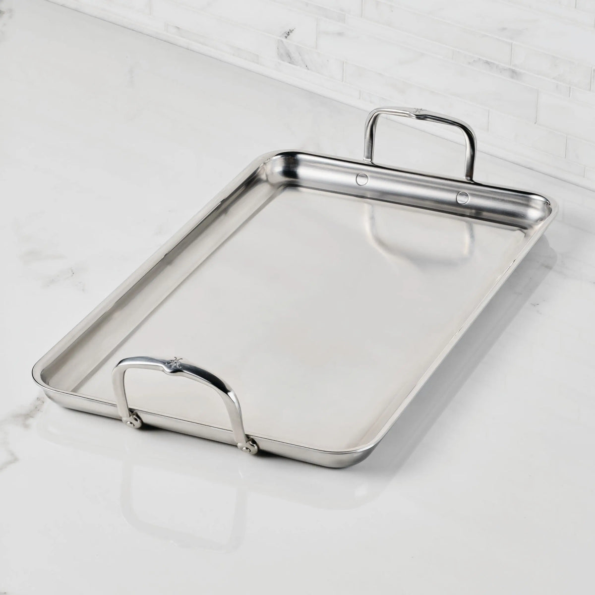 The Hestan Provisions Double Burner Griddle (21x13) features raised edges and two large handles, shown resting on a white marble countertop with a white tiled backsplash in the background.