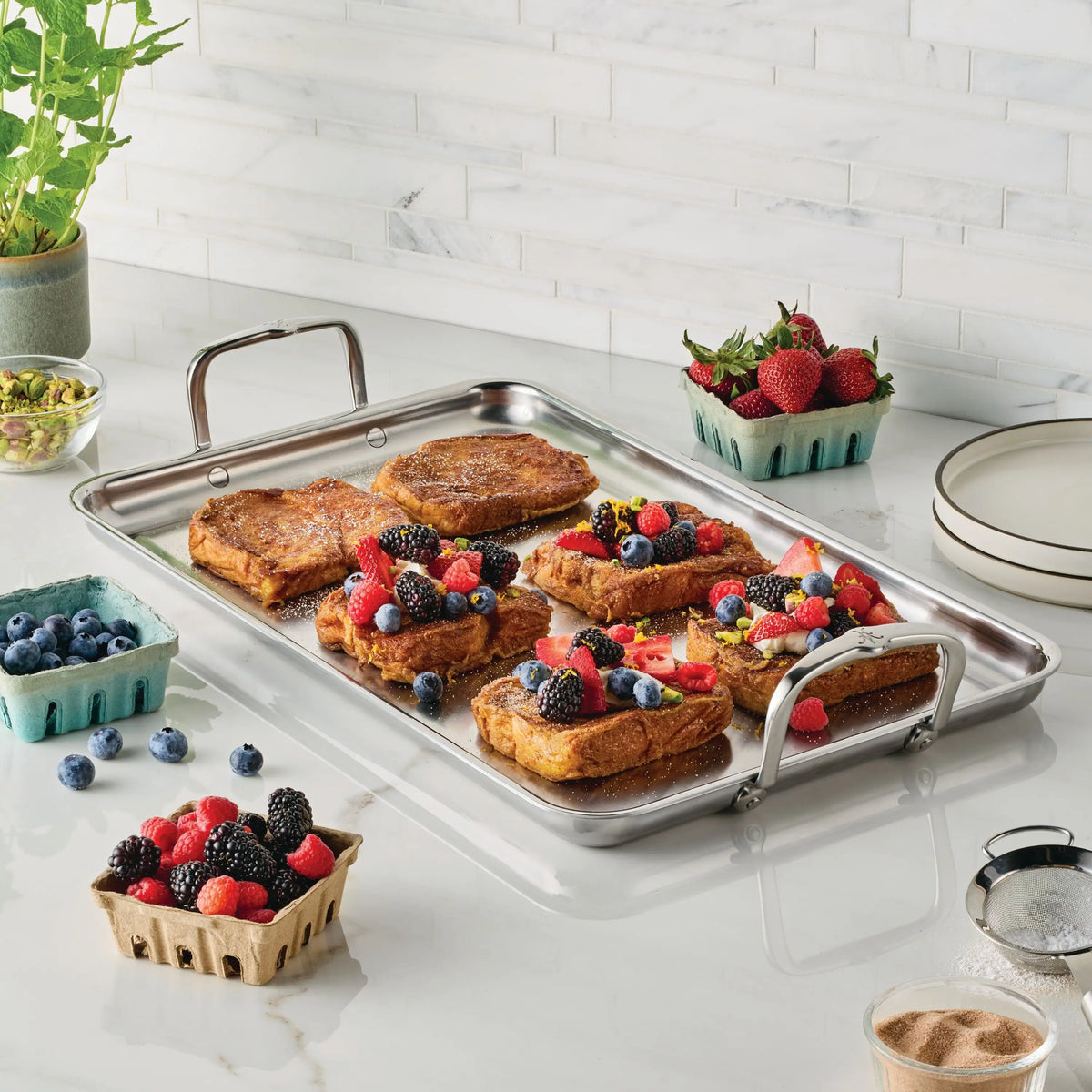A stainless steel tray from the NanoBond Titanium x Tyler Florence Cookware Set, 41-Piece sits on a kitchen counter with French toast topped with mixed berries. Around it are berry cartons, pistachios, potted herbs, and stacked plates.