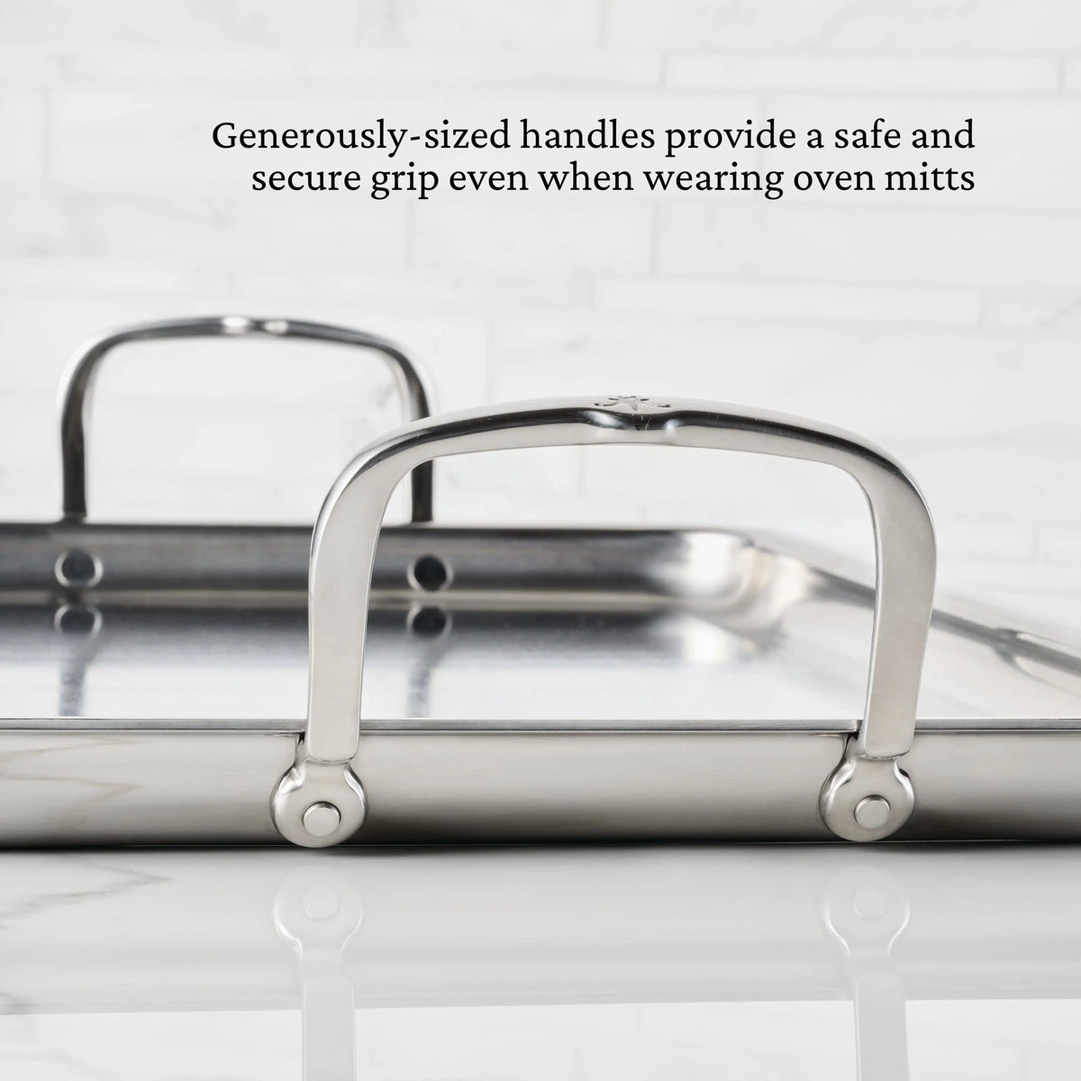 Close-up of the Hestan Provisions Double Burner Griddle (21 x 13) with two large handles for a secure grip, even with oven mitts. Text highlights superb heat distribution. White tiled wall background.