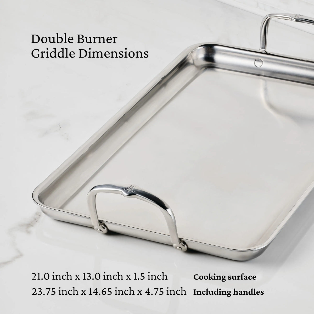 The Hestan Provisions Double Burner Griddle (21 x 13) rests on a white marble surface, highlighting premium stainless steel construction for even heat distribution. Dimensions: 21 x 13 x 1.5 in (cooking), 23.75 x 14.65 x 4.75 in (with handles).